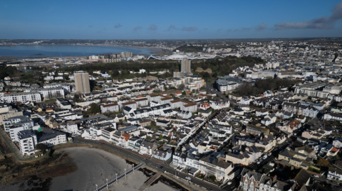 An aerial view of the south of Jersey - rows of houses and a beach in the foreground, sea can also be seen further afield. Blue skies.
