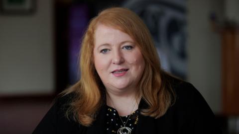 Naomi Long speaking to a reporter at the Stormont Hotel in Belfast in 2024. She has wavy, shoulder-length ginger hair. She is wearing a black cardigan over a black floral patterned top or dress, with a silver necklace which has a pendant made of concentric rings. 