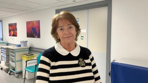 A woman in a black and white stripy jumper stands in a hospital corridor.  She is in her sixties and has medium length brown hair.  In the background are a number of hospital trolleys that carry medical equipment.