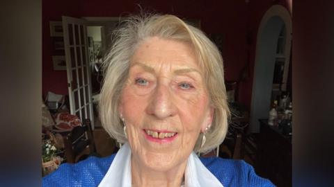 Sylvia Knights pictured in her house. In the background you can see a door open and a drinks shelf and some flowers on a table. Sylvia is wearing a blue jumper and white shirt.