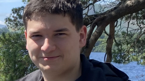 Tom Dingle smiling for a photograph. He has short dark brown/ black hair and is standing in front of a tree and a body of water.