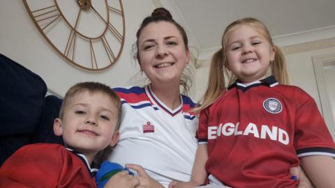 A woman in a white football top sits on a sofa smiling next to her children, wearing red England tops.
