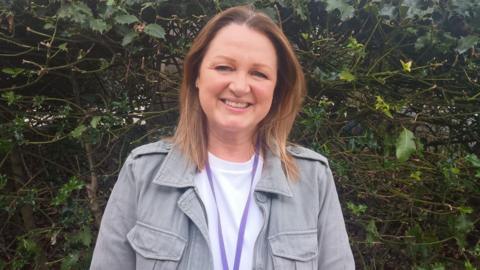 Lisa is standing outside in front of a bush. She has shoulder-length brown hair and is wearing a white top and a khaki jacket. She is smiling at the camera.
