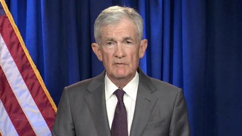 Jerome Powell looks into a camera wearing a grey suit, white shirt and dark striped tie standing in front of a blue curtain and American flag.