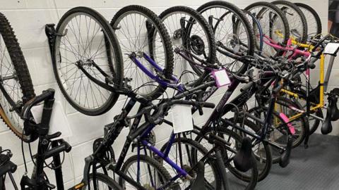 Eight bikes leant up vertically against a white wall. The front wheels of the bikes are touching the wall.
