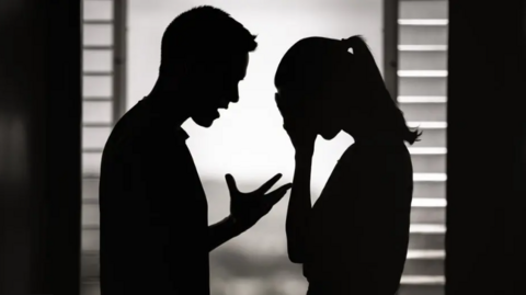 Stock photo of the shadow of a man and a woman facing each other against a window. The man has is mouth open in a talking motion and his hand up in front of him, bent at the elbow. The woman has a ponytail in her hair and her head is in her hands.