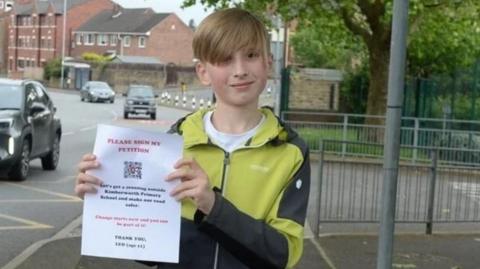 Leo, who has blonde hair and a fringe falling to one side, holds up a piece of paper that reads "please sign my petition". He stands next to a road and three cars can be seen in the background.