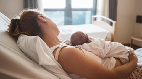 A mother rests in a hospital bed while gently holding her newborn baby. The atmosphere is calm and comforting, showcasing their bond in a serene medical environment.