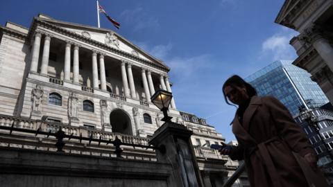 The Bank of England with a blue sky behind it, in the foreground a woman stands on her phone