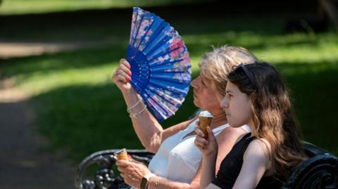 two women sitting on a park bench. One eating an ice cream, the other fanning herself in the hot weather
