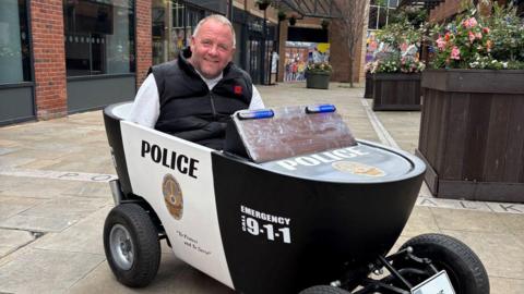 A man sits in a soapbox made up to look like a black-and-white police vehicle.