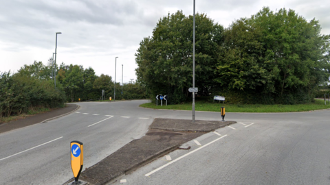A road leading to a roundabout with two green trees in the middle. The sky is grey.