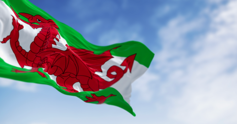 A close-up of the Welsh flag flying in the wind in front of a blue sky. The flag has two green stripes and a white stripe with a red dragon in the middle.