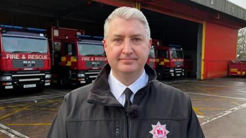 Devon and Somerset Fire Service Gavin Ellis wearing a shirt, tie and a fire service coat. He is stood in front of a fire station with four fire engines inside.