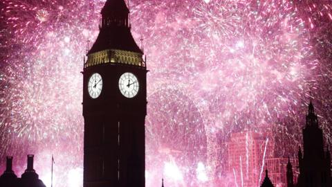 The Elizabeth Tower, better known as Big Ben, is silhouetted against bright pink fireworks exploding against the night sky in  central London, with the London Eye and rooftops in the background. The white clock face on the tower reads 00:11.