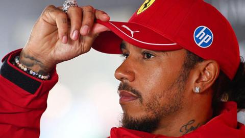 Lewis Hamilton adjusts his cap while speaking to the media before the Dutch Grand Prix. He is wearing a Ferrari cap and coat