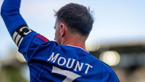 An image of the back of Mason Mount with the stands out of focus in the background. He is wearing the captain's armband and holding his arm in the air.