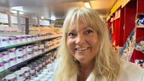 A woman with long blonde hair is smiling at the camera. She is wearing a white lab coat. Behind her are stacks of paint tubs – some of them on shelves.