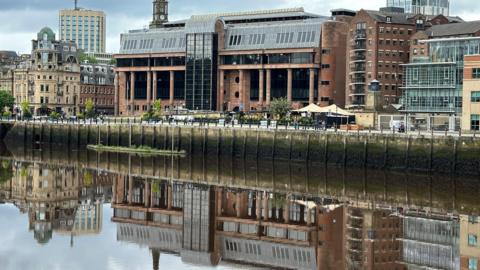 A large court building made of red stone and large dark windows reflected in a river running in front of it