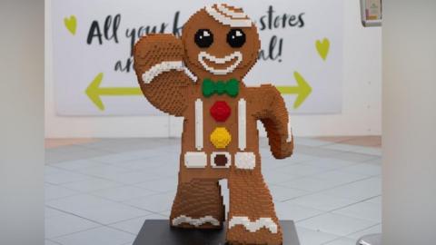 A large figure of a gingerbread man created out of Lego bricks. The colouring is brown with round black eyes, and a white outline of a mouth, and one arm raised. A green bow tie, and a red and yellow button complete the "outfit". It is standing in what appears to be an indoor shopping mall.