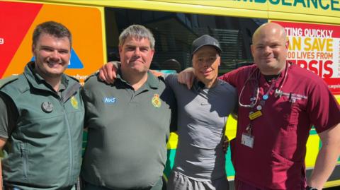 Four men stand smiling in front of an ambulance, two of them wear paramedics' uniforms and one wears scrubs