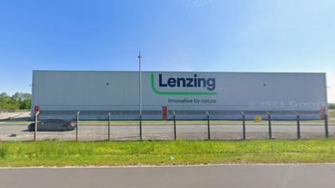 The exterior of Lenzing in Grimsby - a large, boxy, grey building with a large company logo on the front. It is behind a wire fence.