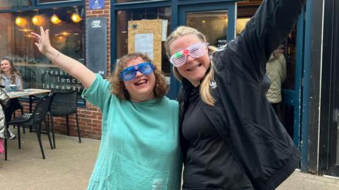 Two women pose outside a coffee shop, with their arm around each other and one hand in the air. The woman on the left has short curly brown hair and wears a mint green top and blue grilled sunglasses. The woman on the right is taller, with long blonde hair in a ponytail, and wears a black t-shirt and black jacket.