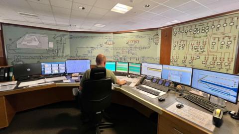 A man sat on a black office chair in a control room with his back to the camera. He is surrounded by nine computer monitors and various control equipment.