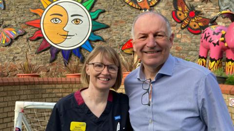 Rachel Hughes and Martin Gargan smile at the camera. Rachel has short brown hair and glasses. She is wearing nurse's scrubs. Martin has short grey hair and is wearing a blue striped shirt with his glasses attached to his shirt. They are standing outside in a kid's play area which is decorated with butterflies, a sun and moon and a Gromit.