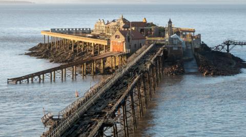 Birnbeck Pier in Weston-super-Mare. The pier is damaged and in disrepair, with a heavily damaged wooden walkway and dilapidated buildings.