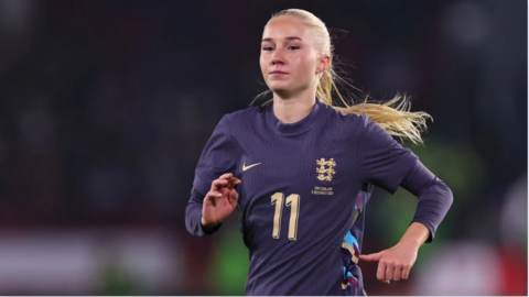 Laura Blindkilde Brown with a blond ponytail and wearing a navy blue England shirt. She is mid-run and is in front of a blurred background.