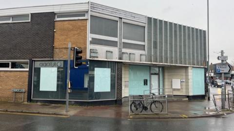 The former Lloyds bank building in Gorleston, featuring black and yellow brick panels, wide windows and a stone-clad facade with vertical separations. The lower floor windows have been obscured and all branding has been removed. Nearby are railings and traffic lights and signs, with a bicycle chained to one of the railings