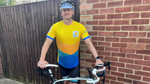 Man wearing a yellow top and shorts, standing next to a bicycle and wearing a safety helmet