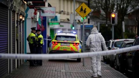 A forensic officer walks towards the restaurant and two uniformed Irish police officers and a police car. There is a cordon at the forefront of the picture