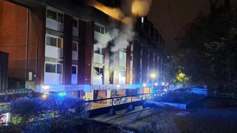 A photo taken in the dark showing a fire engine parked near to the flat where the fire broke out. Several firefighters can be seen stood outside and and a plume of smoke is rising into the air