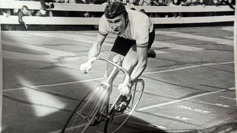 A black and white photo of a man cycling on a track looking strained as crowds cheer him on