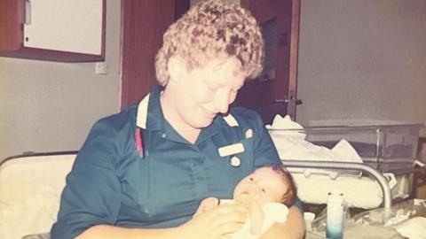 A colour photograph, possibly from the 1980s, of a woman smiling down at a baby she is holding in her arms. She is wearing a dark blue midwife's tunic and has short very curly blonde hair. The baby has dark hair and its eyes are open.
