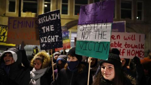 The image shows multiple people wearing winter-appropriate clothing holding signs that read "feminist revolution", "reclaim the night" and "blame the system not the victim". They are standing on a street in the dark.