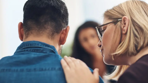 A woman rests her hand on the shoulder of a man with his back to the camera. He is wearing a blue shirt and has dark hair. She has short blonde hair and glasses on.
