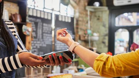 Young woman wearing a yellow top with rolled-up sleeves pays for goods with a smart watch in a coffee shop.