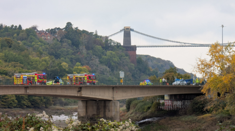 Two fire engines and two police cars are blocking the road on a concrete bridge. Their blue lights are on. A blue screen has been put up while emergency services work and a silver car can be seen on one side of the bridge. In the background are trees on the side of Avon Gorge and part of Clifton Suspension Bridge is visible in the distance.