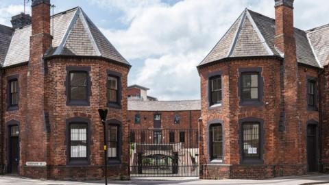 A view of a horse-shoe shaped 18th century brick building with black framed windows and black iron gates at the front 