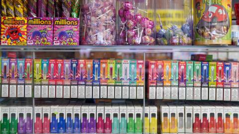 Disposable vapes are on display in a glass cabinet across two shelves. They are all different colours: pink, green, purple, yellow, blue, orange, red and white. On top of the glass cabinet are sweets. On the left are tubes of millions in orange and pink packaging and on the right are four boxes of lollipops. Some are bubble-gum pink whilst others are blue, green and red.