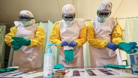 Three health workers stand side-by-side as they put on full PPE. They are wearing goggles and protected by yellow and white overalls and are pictured putting on green gloves. 