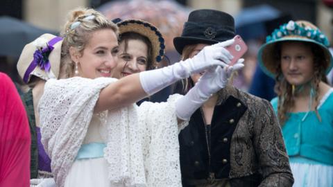 Two young women dressed in outfits from Jane Austen's era smile as they take a selfie during the Jane Austen Festival in Bath. The one most in shot is wearing a white dress with a light blue belt, and has pearl-style earrings. In the background other women in similar outfits are visible