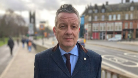 Nick Kemp, with short grey hair is standing on a high street. he is wearing a blue suit and is looking at the camera, expressionless.