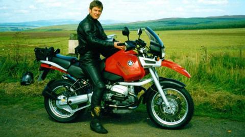 Gordon Castle is sitting on a large BMW bike wearing motorbike leathers. Behind him is a large stretch of hills and moorland under a grey and blue sky. A helmet is on the grass behind.
