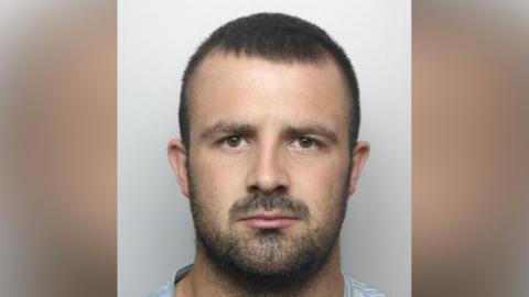 A police mugshot of Ross Daniels. He has short brown hair and dark brown facial hair.