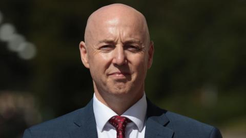 A man, Jon Burrows, is staring at the camera. He is bald and is wearing a dark suit jacket over a white shirt and red tie. He has a neutral expression on his face. He is standing outside, the out-of-focus background appears to be trees. 
