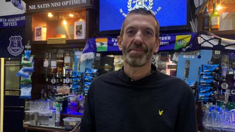 Dave stands in front of an adorned bar filled with Everton scarves. He is wearing a dark polo shirt and has short dark hair